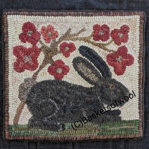 May include: Hand-hooked wool rug featuring a black rabbit beneath a flowering tree with red blossoms. The rug has a neutral background with a dark border and green grass at the bottom. The design is a square shape.