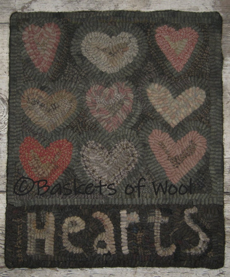 Op de afbeelding: Handgemaakt tapijt met negen hartvormen in rood, roze en grijs op een donkere achtergrond. Het woord "Hearts" staat onderaan. Het tapijt is gemaakt van wol.