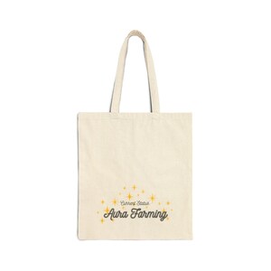 May include: Cream-colored tote bag with the text "Current Status" and "Aura Farming" in a stylized font. The design includes gold starbursts. The bag has two long handles for carrying.