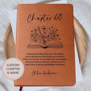 May include: A tan leather-bound journal featuring a black line art illustration of an open book with flowers growing from the pages. The text reads Chapter 60, followed by a paragraph about life and the name Chloe Anderson. A circular label says CUSTOM CHAPTER & NAME.