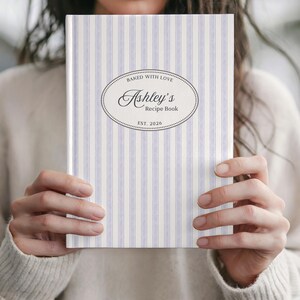 May include: A personalized recipe book with a light blue and white striped cover. The cover features an oval design with the text "Baked with Love" and "Ashley's Recipe Book" with the establishment year 2026.