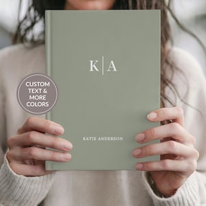 May include: Sage green journal with white text. The cover features the initials "KA" and the name "Katie Anderson." A circular graphic in the corner reads "CUSTOM TEXT & MORE COLORS."
