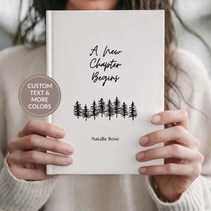 May include: A white hardcover book featuring the black text A New Chapter Begins, a black graphic of a row of pine trees, and the name Natalie Rose printed at the bottom. A circular badge on the left displays the text CUSTOM TEXT & MORE COLORS.