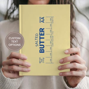 May include: A pale yellow hardcover journal featuring a dark blue graphic design that mimics a stick of salted butter. The cover includes text reading "SALTED BUTTER", "GRADE AA", "NET WT. 4 OZ. (113 g)", and a measurement scale with tablespoons and cups.