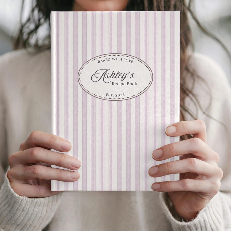 May include: A recipe book with a vertical striped pattern in white and light purple. The cover features an oval design with the text "Baked with Love", "Ashley's Recipe Book", and "EST. 2026".