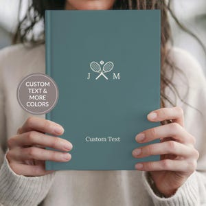 May include: Teal-colored notebook with a tennis racket and ball graphic. The notebook has the initials "J" and "M" and the words "Custom Text" printed on the cover. The notebook is held open.