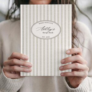 May include: A personalized recipe book with a striped pattern in shades of beige and white. The cover features an oval design with the text "Baked with Love" and "Ashley's Recipe Book, Est. 2026."