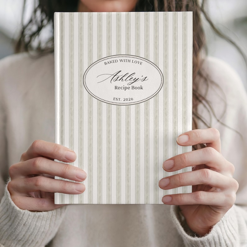 May include: A personalized recipe book with a striped pattern in shades of beige and white. The cover features an oval design with the text "Baked with Love" and "Ashley's Recipe Book, Est. 2026."