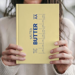 Puede incluir: Un cuaderno amarillo claro con la inscripci&oacute;n "SALTED BUTTER" en azul. Incluye una gu&iacute;a de medidas en cucharadas y tazas. El peso neto es de 113 g. El cuaderno est&aacute; sostenido por una persona.