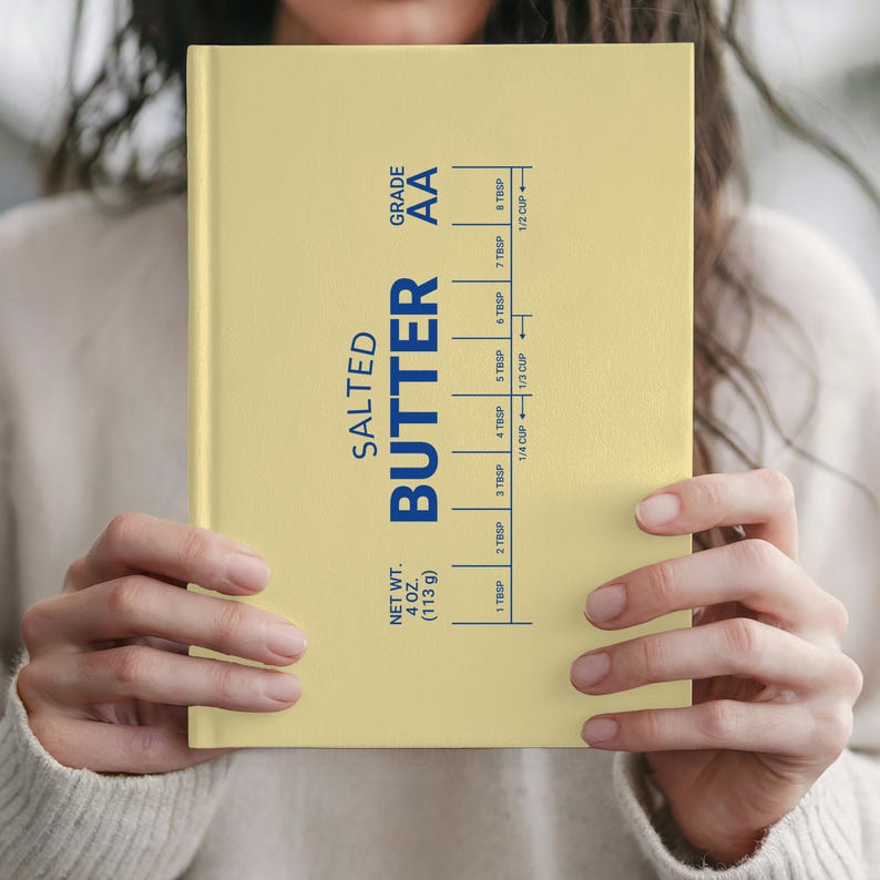 Puede incluir: Un cuaderno amarillo claro con la inscripci&oacute;n "SALTED BUTTER" en azul. Incluye una gu&iacute;a de medidas en cucharadas y tazas. El peso neto es de 113 g. El cuaderno est&aacute; sostenido por una persona.