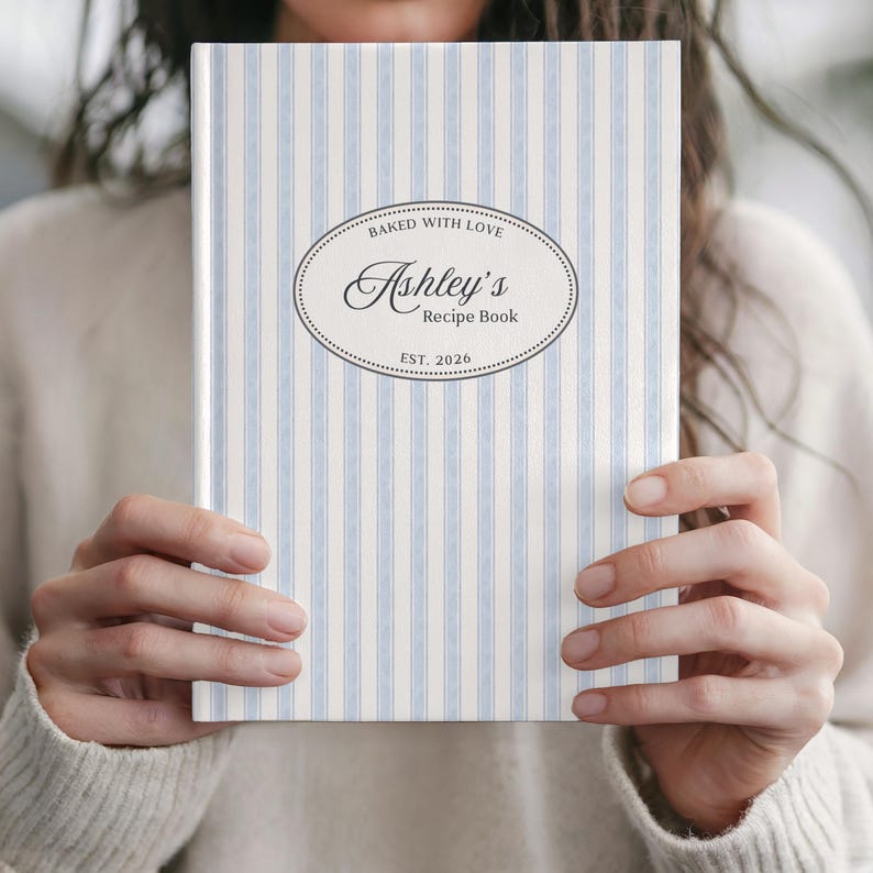 May include: A personalized recipe book with a light blue and white striped cover. The cover features an oval design with the text "Baked with Love" and "Ashley's Recipe Book" with "EST. 2026". The book is held by a person.