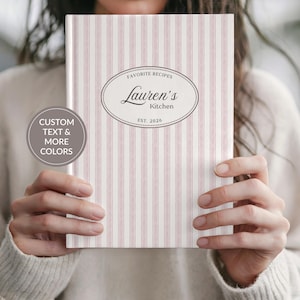 May include: A recipe book with a pink and white striped cover. The cover features an oval design with the text "Favorite Recipes" above "Lauren's Kitchen" and "EST. 2026". A circular graphic on the left says "CUSTOM TEXT & MORE COLORS".