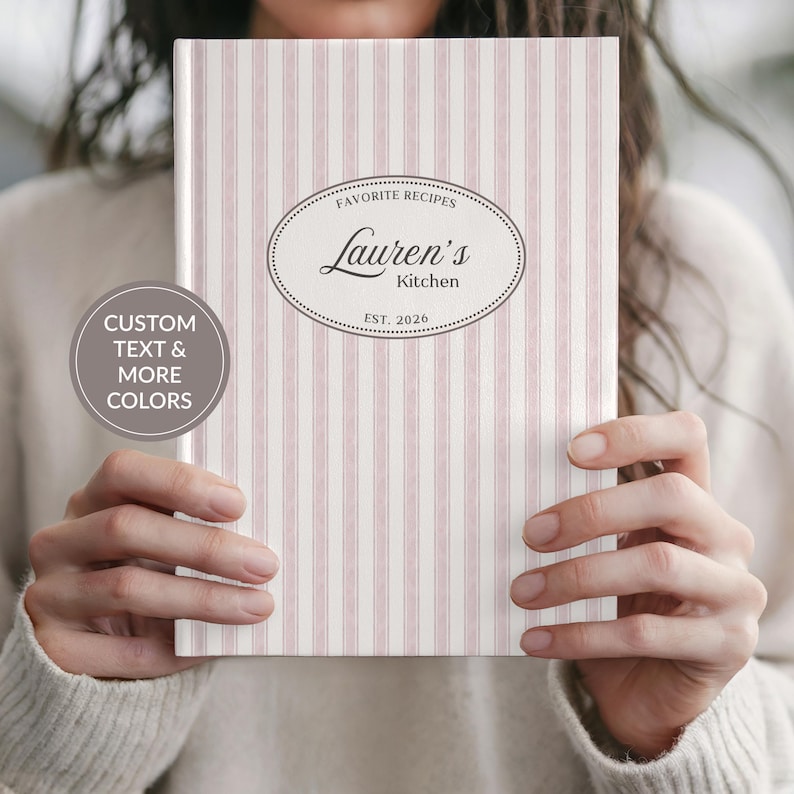 May include: A recipe book with a pink and white striped cover. The cover features an oval design with the text "Favorite Recipes" above "Lauren's Kitchen" and "EST. 2026". A circular graphic on the left says "CUSTOM TEXT & MORE COLORS".