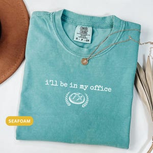 May include: Seafoam green t-shirt with the text "I'll be in my office" in white, and a graphic. A gold necklace with a pendant rests on the shirt. The shirt is folded, and a brown hat is partially visible.