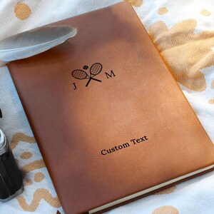 May include: A brown leather-like journal featuring a black graphic of two crossed tennis rackets with a tennis ball and the letters J and M. The cover also includes the printed text Custom Text in a black serif font centered near the bottom.