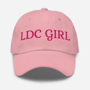 Puede incluir: Gorra de béisbol rosa claro con el texto bordado "LDC GIRL" en rosa más oscuro. La gorra tiene una visera curva y una corona redondeada, típica de las gorras de béisbol.