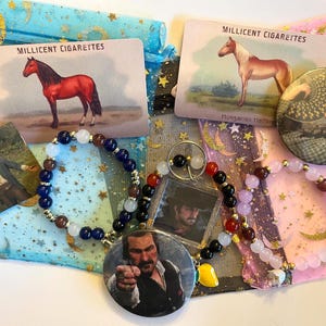 Op de afbeelding: Een verzameling items in vintage-stijl, waaronder sigarettenkaarten met paarden, armbanden met verschillende kralenkleuren en karakter-speldjes. De items zijn gerangschikt op decoratieve stoffen zakjes met ster- en maanpatronen.