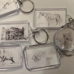 May include: A selection of clear acrylic keyrings featuring monochrome sketches. Designs include a horse, a deer, a house, flowers, and a cross. Each keyring has a silver metal ring.