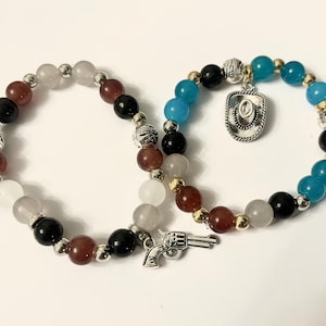 May include: Two beaded bracelets. One features red, black, white, and silver beads. The other has blue, black, and gold beads, with a silver cowboy hat charm and a silver gun charm. The bracelets are on a white surface.