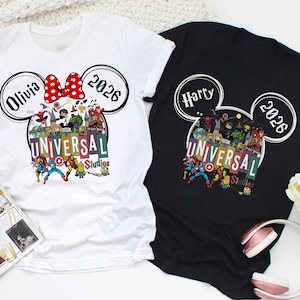 Peut inclure: Deux t-shirts, un blanc et un noir, avec un design en forme de Mickey Mouse. Le t-shirt blanc porte le nom "Olivia" et l'année "2026" avec un nœud rouge. Le t-shirt noir affiche le nom "Harry" et l'année "2026". Les deux t-shirts présentent un graphisme "Universal Studios".