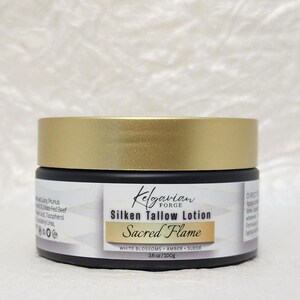 Può includere: Un barattolo nero con coperchio dorato, etichettato "Kelgavian Forge Silken Tallow Lotion Sacred Flame". L'etichetta elenca gli ingredienti e le dimensioni del prodotto come 100g. La lozione è per la cura della pelle.