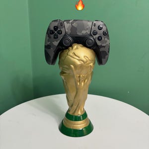 May include: A gold trophy-shaped PS5 controller holder with a green base. A camouflage-patterned controller rests on top. The text "World Cup PS5 holder" is displayed above the controller.