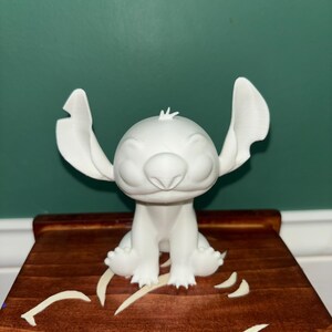 Puede incluir: Una figura blanca impresa en 3D del personaje Stitch de la pel&iacute;cula de Disney Lilo & Stitch. La figura est&aacute; sentada con los brazos a los lados y las orejas grandes extendidas. La figura est&aacute; sobre una superficie de madera.