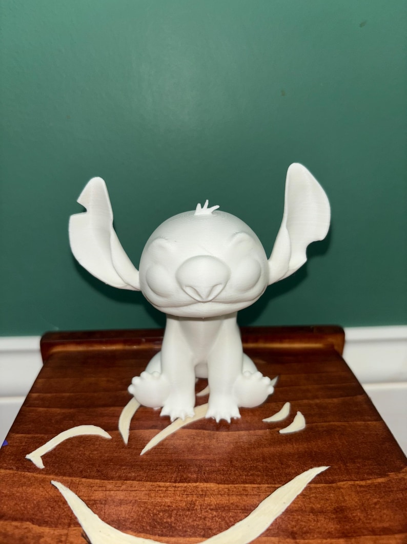 Puede incluir: Una figura blanca impresa en 3D del personaje Stitch de la pel&iacute;cula de Disney Lilo & Stitch. La figura est&aacute; sentada con los brazos a los lados y las orejas grandes extendidas. La figura est&aacute; sobre una superficie de madera.