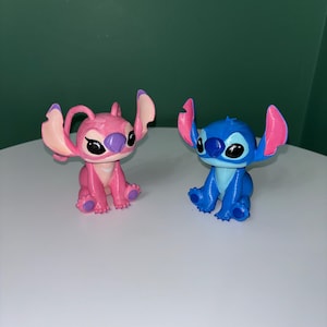 Puede incluir: Dos figuras de los personajes Stitch y Angel de la pel&iacute;cula animada Lilo & Stitch. La figura azul de Stitch tiene orejas grandes y una amplia sonrisa. La figura rosa de Angel tiene caracter&iacute;sticas similares. Ambos est&aacute;n sentados sobre una superficie blanca.