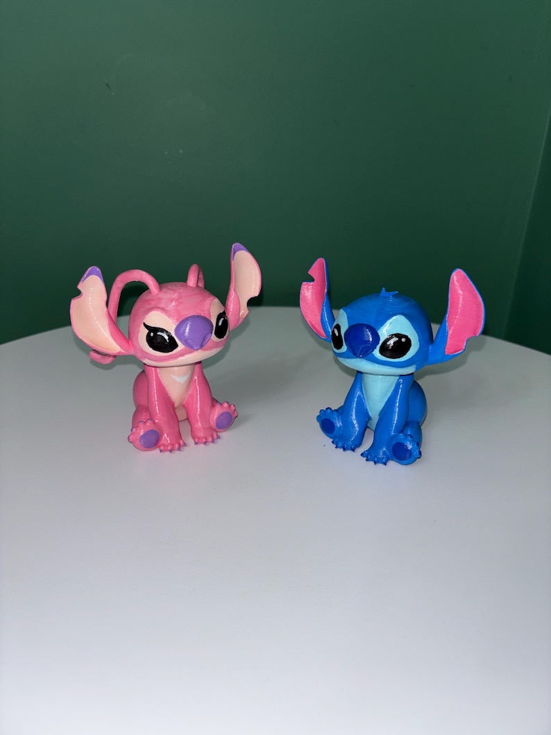 Puede incluir: Dos figuras de los personajes Stitch y Angel de la pel&iacute;cula animada Lilo & Stitch. La figura azul de Stitch tiene orejas grandes y una amplia sonrisa. La figura rosa de Angel tiene caracter&iacute;sticas similares. Ambos est&aacute;n sentados sobre una superficie blanca.