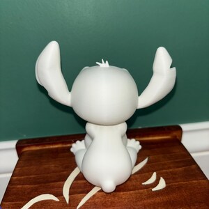 Puede incluir: Una figura blanca de un personaje de dibujos animados con orejas grandes y una cola peque&ntilde;a, sentada sobre una superficie de madera. La figura es lisa y tiene un dise&ntilde;o sencillo. La superficie de madera est&aacute; te&ntilde;ida de marr&oacute;n con detalles decorativos blancos.
