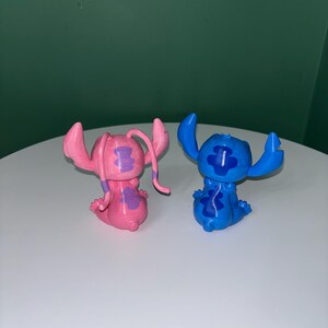 Puede incluir: Dos peque&ntilde;as figuras del personaje Stitch de la pel&iacute;cula animada Lilo & Stitch. Una es rosa con detalles morados, y la otra es azul con detalles azul oscuro. Ambas posan con los brazos en alto y tienen orejas grandes.