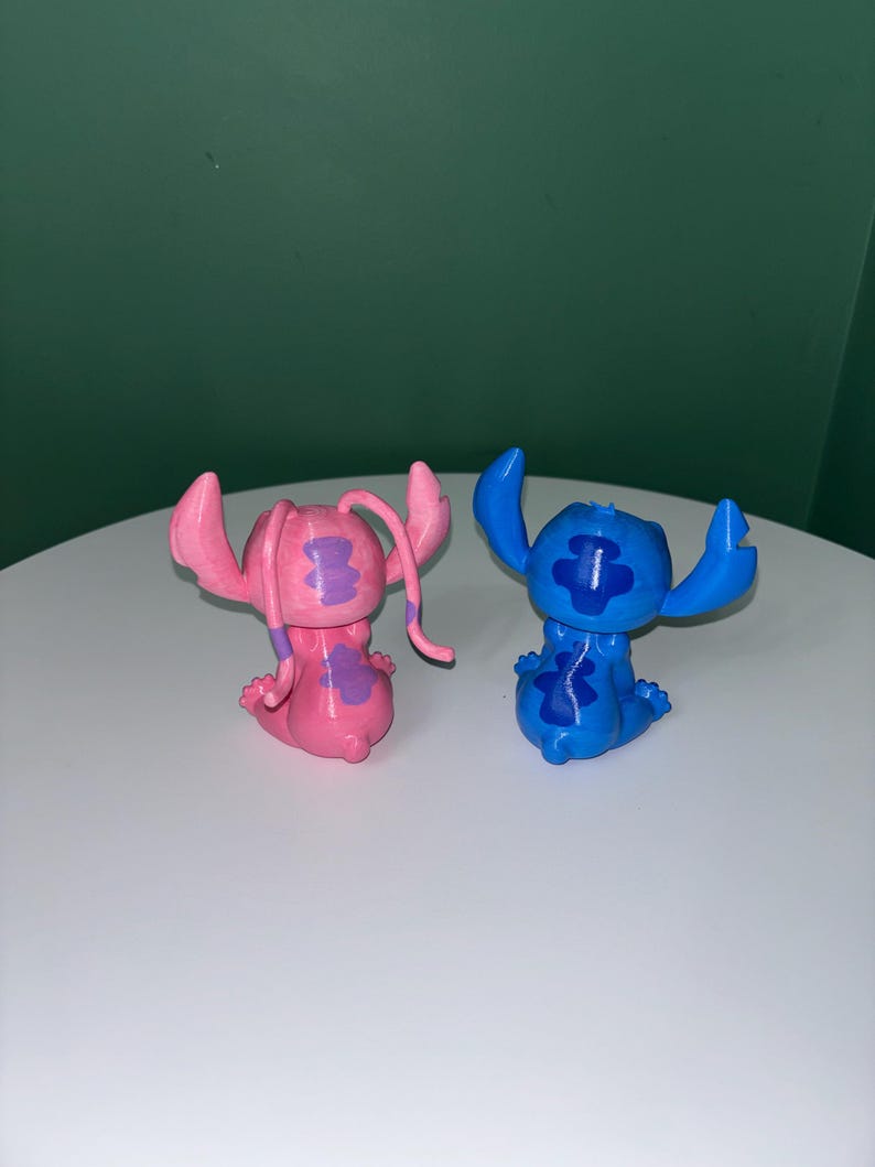 Puede incluir: Dos peque&ntilde;as figuras del personaje Stitch de la pel&iacute;cula animada Lilo & Stitch. Una es rosa con detalles morados, y la otra es azul con detalles azul oscuro. Ambas posan con los brazos en alto y tienen orejas grandes.
