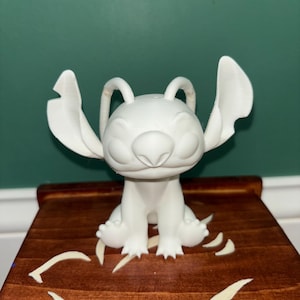Puede incluir: Una figura blanca de un personaje de dibujos animados con orejas grandes y una amplia sonrisa. La figura est&aacute; sobre una superficie de madera con un dise&ntilde;o decorativo. El fondo es de color verde liso.
