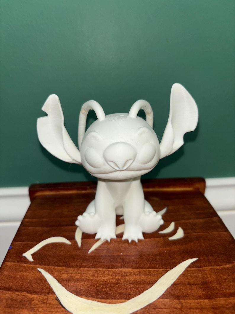 Puede incluir: Una figura blanca de un personaje de dibujos animados con orejas grandes y una amplia sonrisa. La figura est&aacute; sobre una superficie de madera con un dise&ntilde;o decorativo. El fondo es de color verde liso.