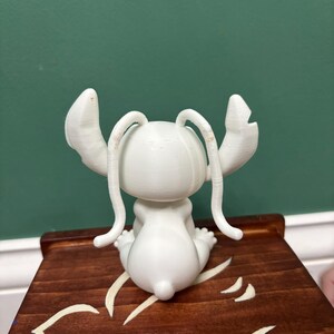 Puede incluir: Una figura blanca de un personaje de dibujos animados con orejas grandes y antenas, sentada sobre una superficie de madera. La figura est&aacute; detallada con texturas visibles. La superficie de madera tiene un acabado marr&oacute;n oscuro con un dise&ntilde;o decorativo de color claro.