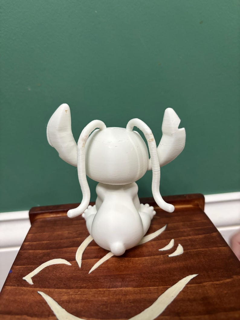 Puede incluir: Una figura blanca de un personaje de dibujos animados con orejas grandes y antenas, sentada sobre una superficie de madera. La figura est&aacute; detallada con texturas visibles. La superficie de madera tiene un acabado marr&oacute;n oscuro con un dise&ntilde;o decorativo de color claro.