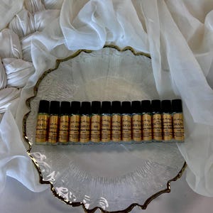 May include: A collection of perfume roller bottles with gold caps and labels, arranged on a decorative glass plate with a gold rim. The labels include text such as "VANILLA", "CHO", and "YALA BRAZIL". The bottles are displayed on a white fabric background.