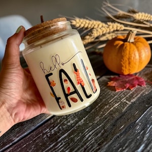 May include: A clear glass candle jar with a cork lid, filled with off-white wax. The front of the jar features black text that reads hello FALL, decorated with small colorful illustrations of autumn leaves, a scarf, and a coffee cup.