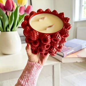 May include: A red candle in a decorative container shaped like raspberries. The candle has a creamy white wax and two wooden wicks. The container is being held up by a person. A vase of tulips is in the background.