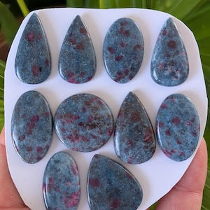 May include: A collection of ten polished gemstones with a blue-gray base color and red inclusions. The stones are various shapes, including teardrop, oval, and round, and are arranged on a white surface. The gemstones are likely for jewelry making.