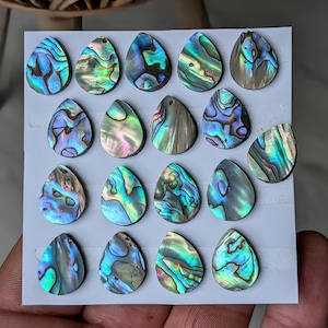 May include: A selection of teardrop-shaped abalone shell pendants. The iridescent shells display blues, greens, and purples. Each pendant has a unique pattern and is mounted on a grey card.