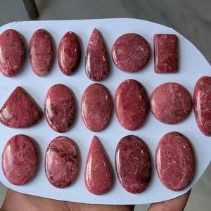 May include: A collection of polished rhodonite gemstones in various shapes, including ovals, teardrops, and rectangles. The stones display a deep pink hue with dark veining, arranged on a white surface.