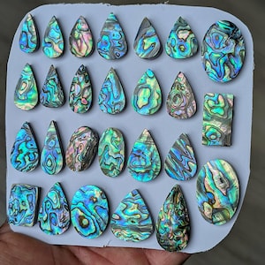 May include: A collection of iridescent abalone shell cabochons in various shapes, including teardrops, ovals, and rectangles. The shells display a vibrant mix of blues, greens, and purples, with unique patterns. These polished shells are ready for jewelry making or crafting.