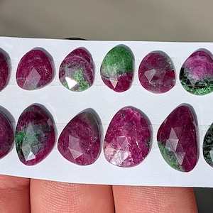 May include: A collection of faceted ruby gemstones with green inclusions, arranged on a white card. The rubies display a deep red hue with varying patterns of green, showcasing their natural beauty. These gemstones are ideal for jewelry making.