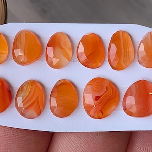 May include: Eleven polished carnelian gemstones, oval in shape, in shades of orange and red. Some stones show natural banding. Each is faceted, reflecting light. The gemstones are displayed on a white card. The stones are suitable for jewellery making or collecting.