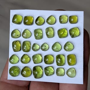 Puede incluir: Una colección de veinticuatro piedras preciosas de color verde oliva de varias formas, incluyendo cuadrados, óvalos y formas redondeadas, están dispuestas sobre una tarjeta blanca. Las piedras tienen un aspecto brillante y transparente, probablemente peridoto.