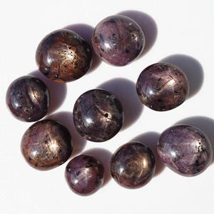 May include: A collection of ten polished, round gemstones. The stones have a deep purple hue with a star-like effect and a metallic sheen. The gems vary slightly in size and are arranged on a white surface.
