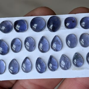 Peut inclure: Une collection de pierres précieuses polies, bleu foncé, de formes variées, dont ovales, en goutte et carrées. Les gemmes sont disposées sur une carte blanche, mettant en valeur leurs surfaces réfléchissantes et leurs motifs internes. Le thème général est la création de bijoux.