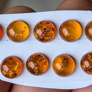 Puede incluir: Nueve cabujones de ámbar redondos y pulidos dispuestos en dos filas sobre un soporte blanco. Cada piedra preciosa muestra un tono naranja amarillento cálido y translúcido con inclusiones internas visibles y motas doradas naturales.